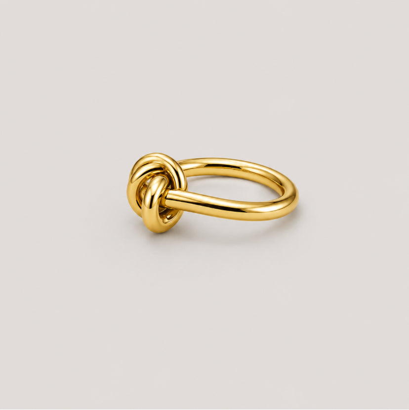 A polished gold ring featuring a knotted loop design on a smooth, tapered band, set against a solid light gray background.