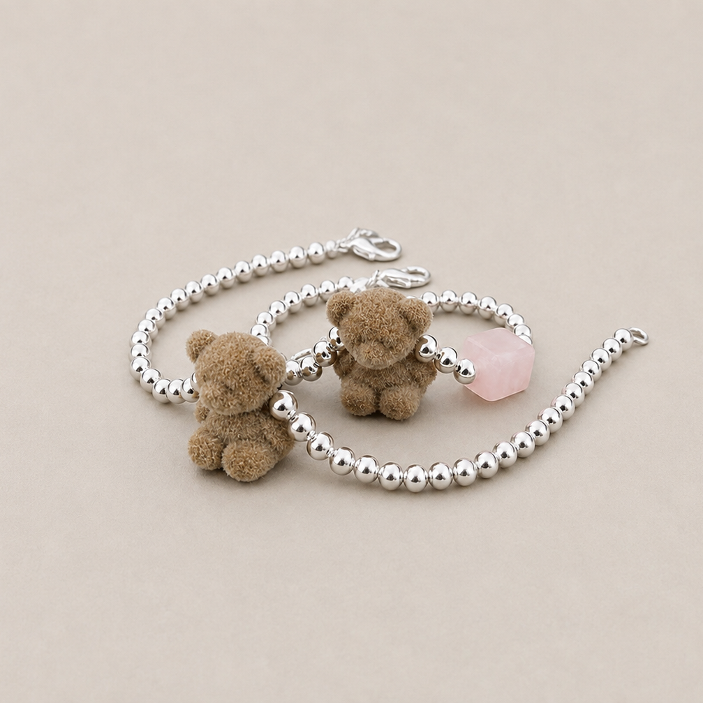 Two silver beaded bracelets with small brown teddy bear charms and a pink stone accent, displayed on a neutral beige background.