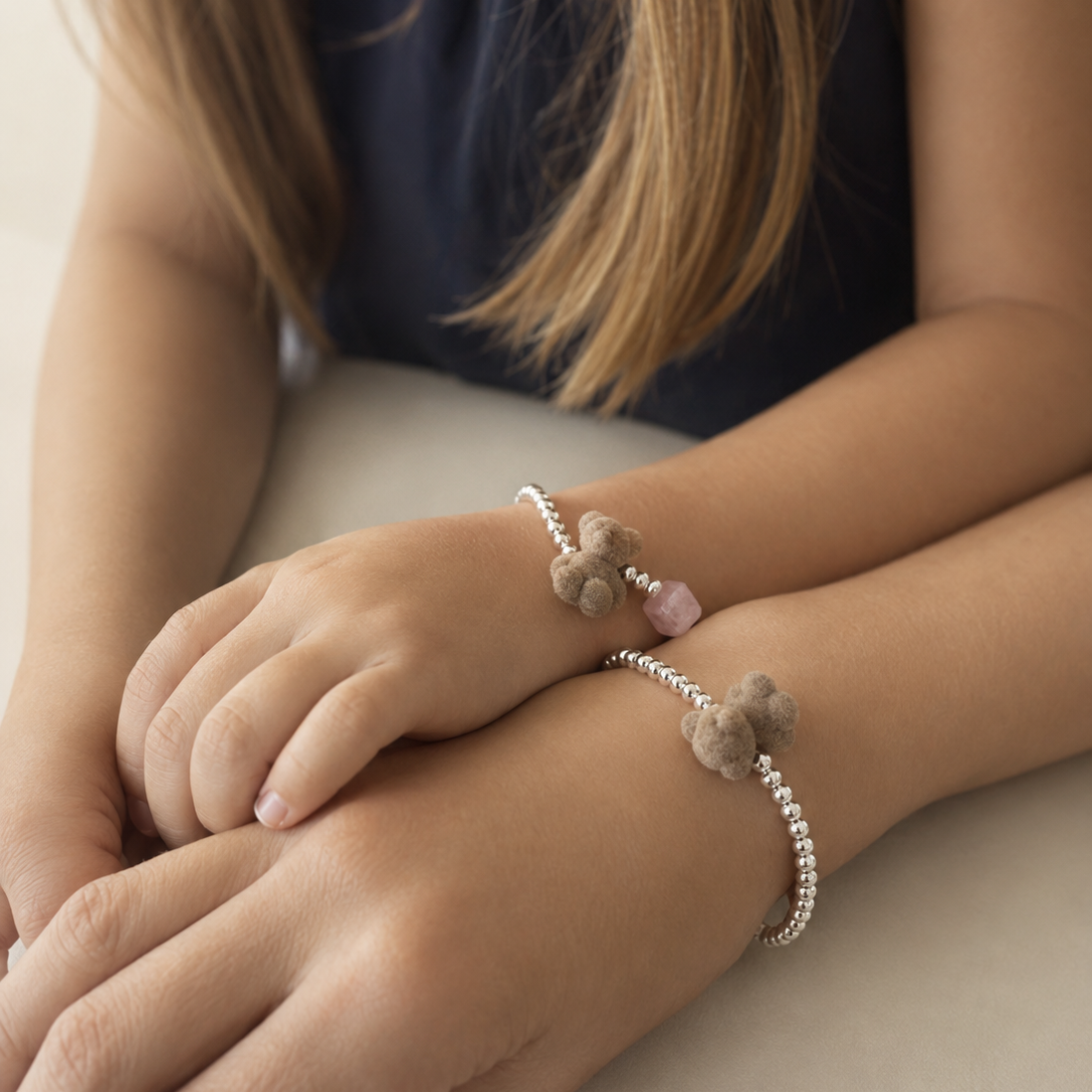 Two delicate beaded bracelets with small gray pom-pom charms and a pink bead, worn on a child's wrists. The child is wearing a dark navy top, and the background is a soft, neutral beige surface.