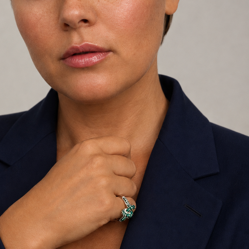 A close-up portrait of a woman wearing a dark navy blazer and a prominent silver ring with a green stone, set against a neutral gray background.