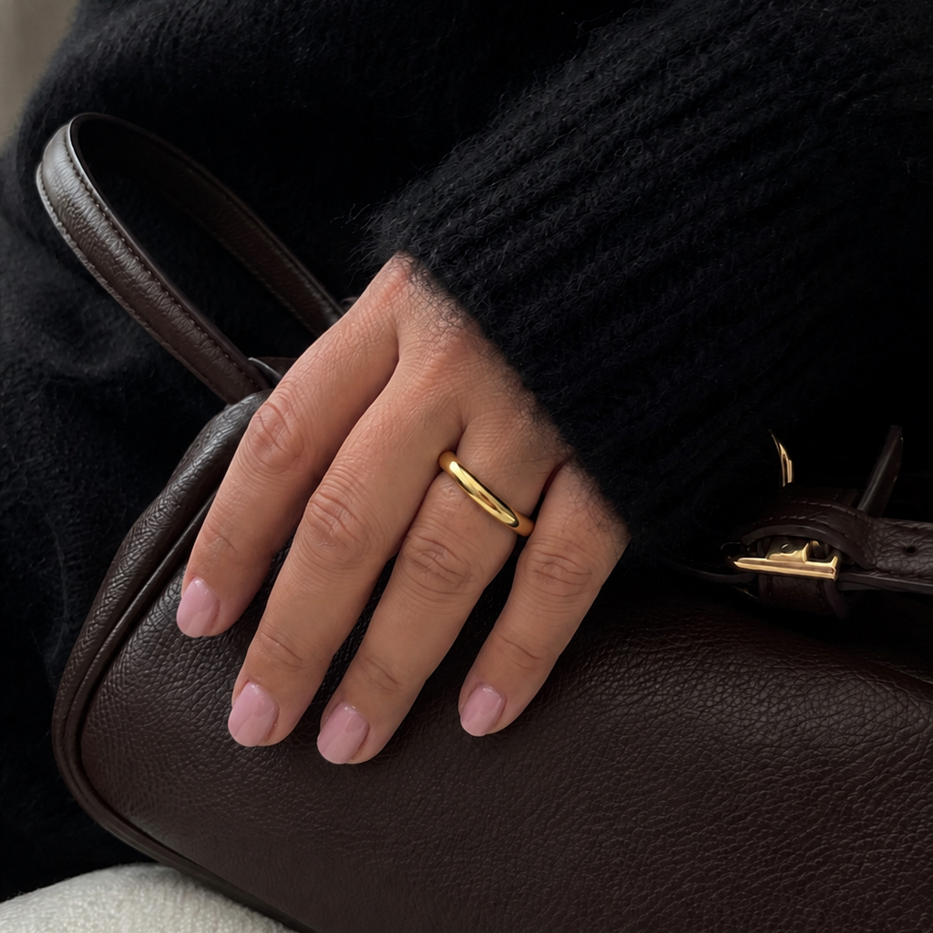 A close-up of a person's hand resting on a dark brown textured leather handbag, wearing a simple gold band ring. The person is dressed in a black ribbed knit sweater.