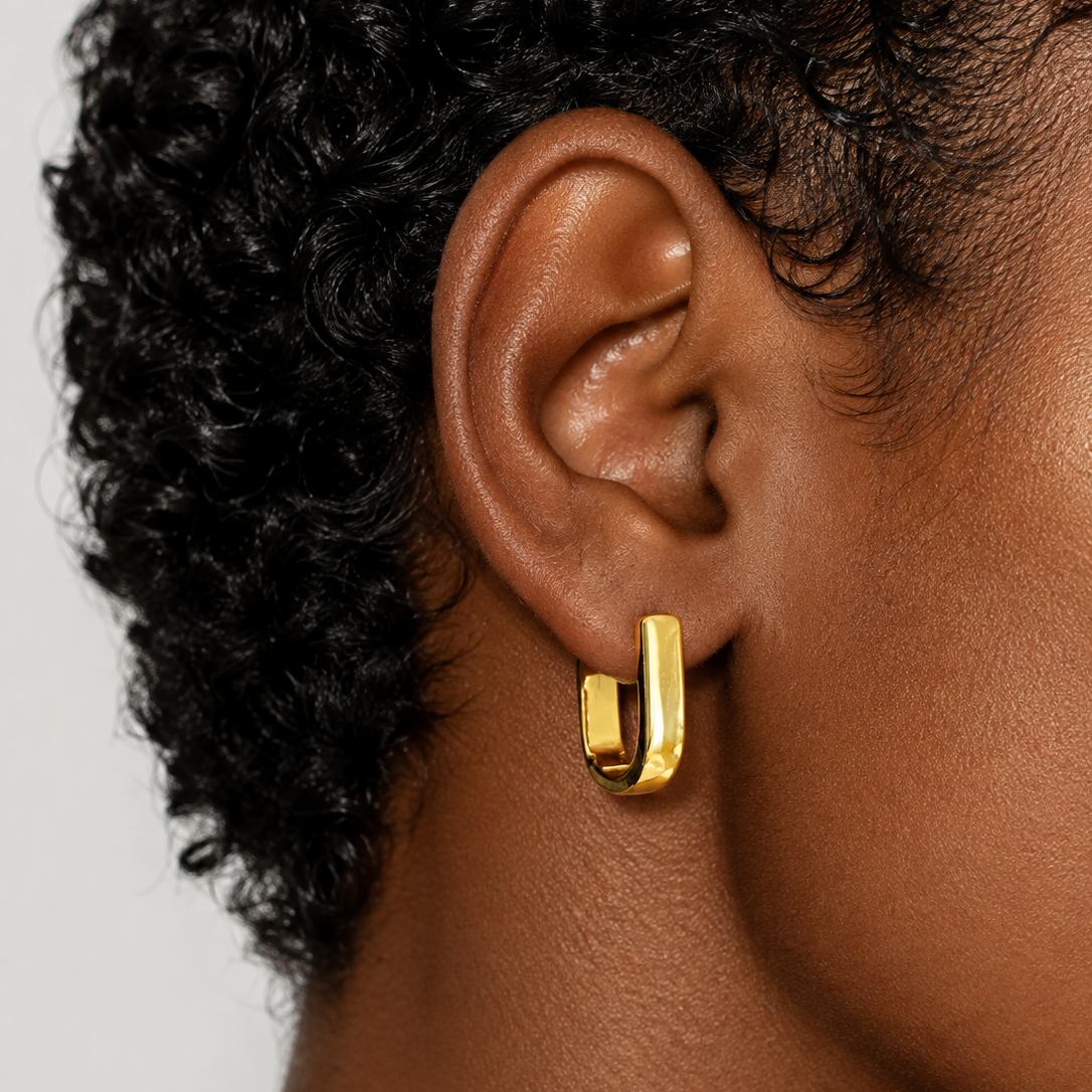 A close-up of a person's ear wearing a small gold huggie hoop earring against a light gray background.