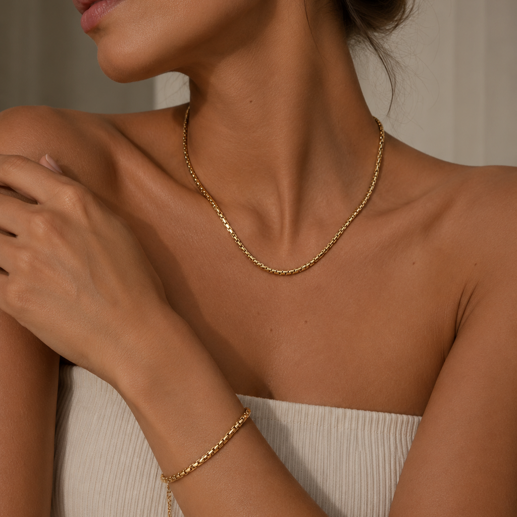 A close-up lifestyle shot of a model wearing a delicate gold chain necklace and a matching gold chain bracelet, paired with a cream-colored ribbed strap top.