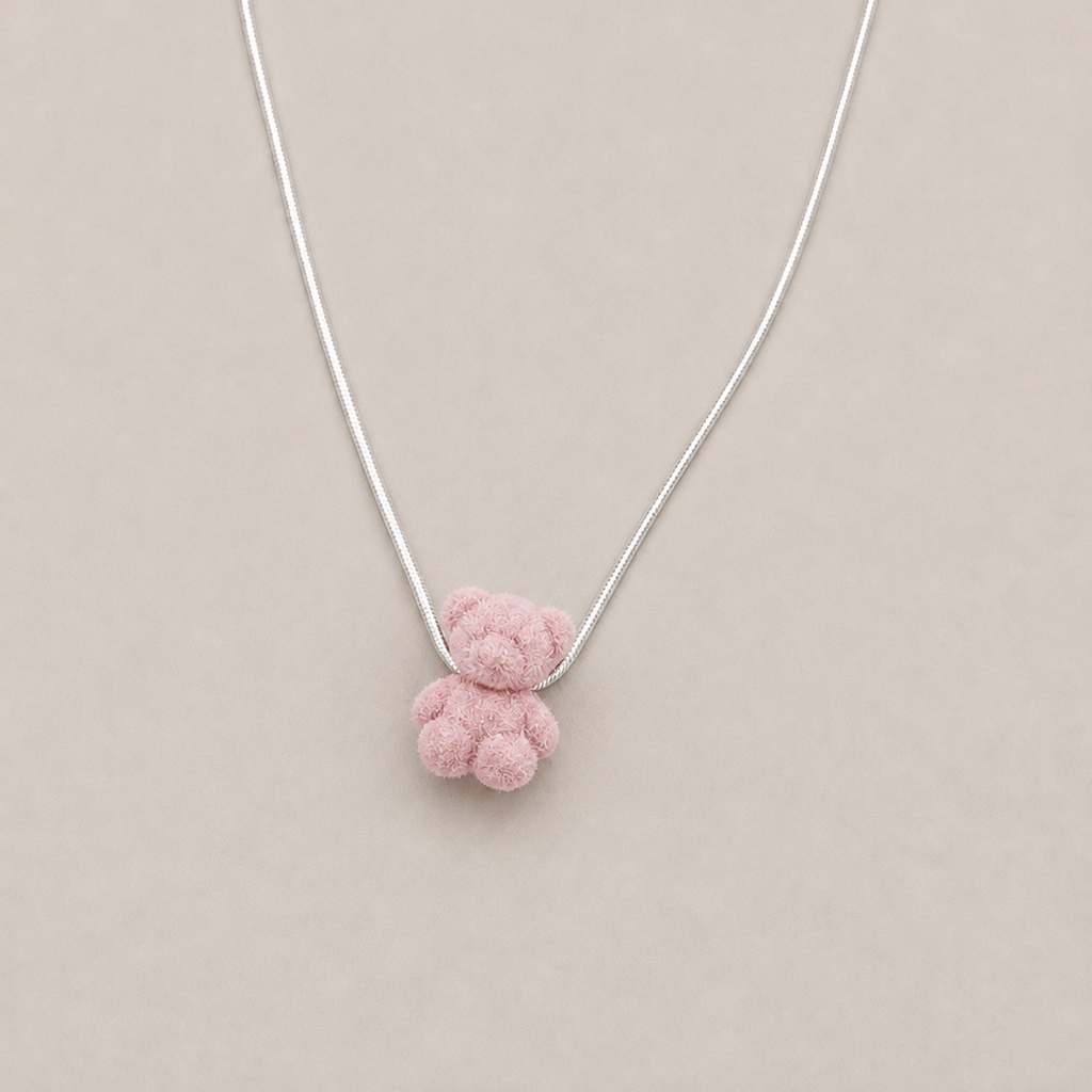 A delicate silver chain necklace featuring a small, soft pink textured teddy bear pendant, displayed against a solid light beige background.