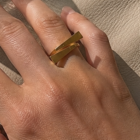 A close-up of a person's hand resting on a beige leather surface, wearing a chunky gold ring. A ribbed clear glass tumbler is partially visible in the upper left, casting a shadow across the hand and leather background.