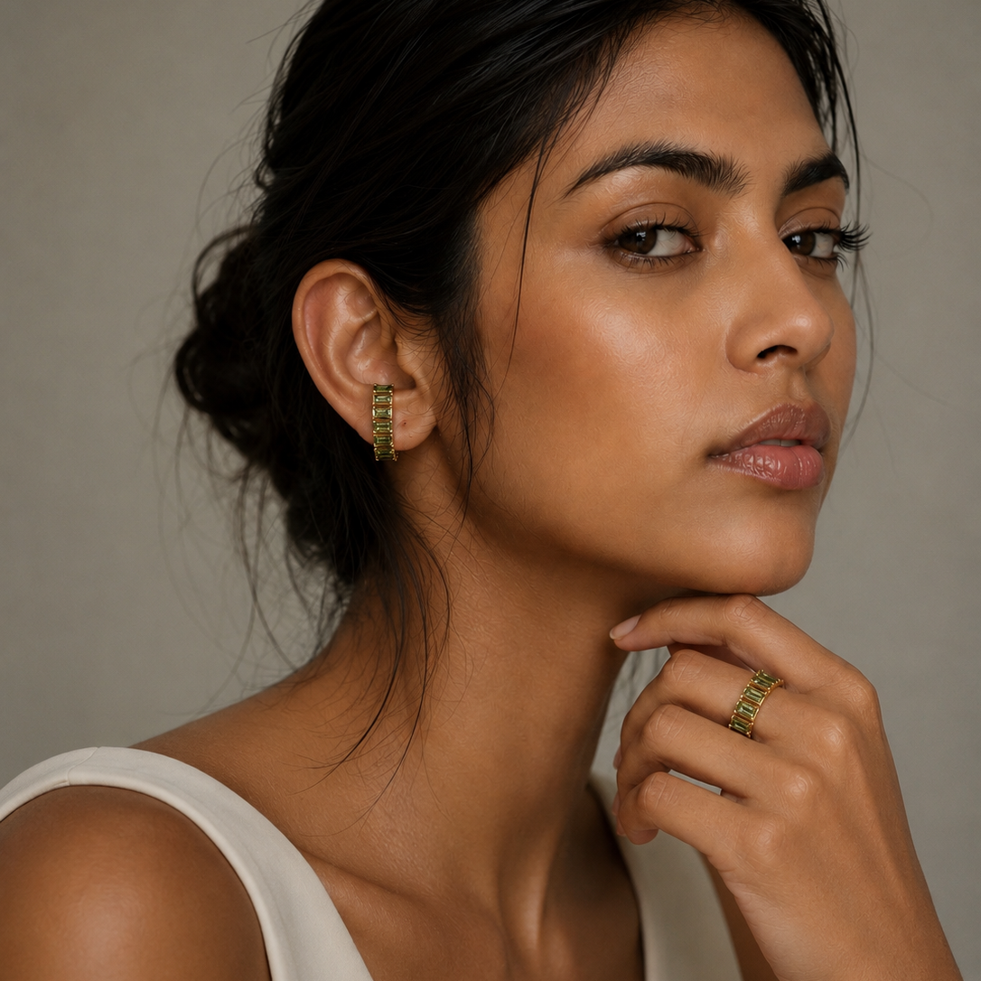 A close-up portrait of a woman wearing small gold hoop earrings and a gold ring, with her hand resting near her chin against a neutral gray background.