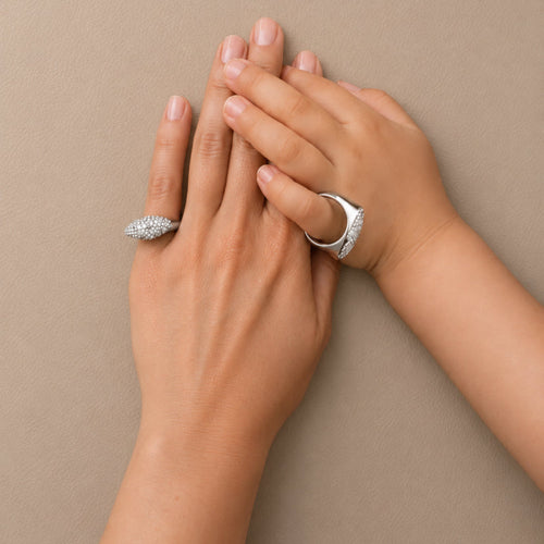 Two hands resting against a neutral beige background, each wearing a silver ring with clear gemstones.