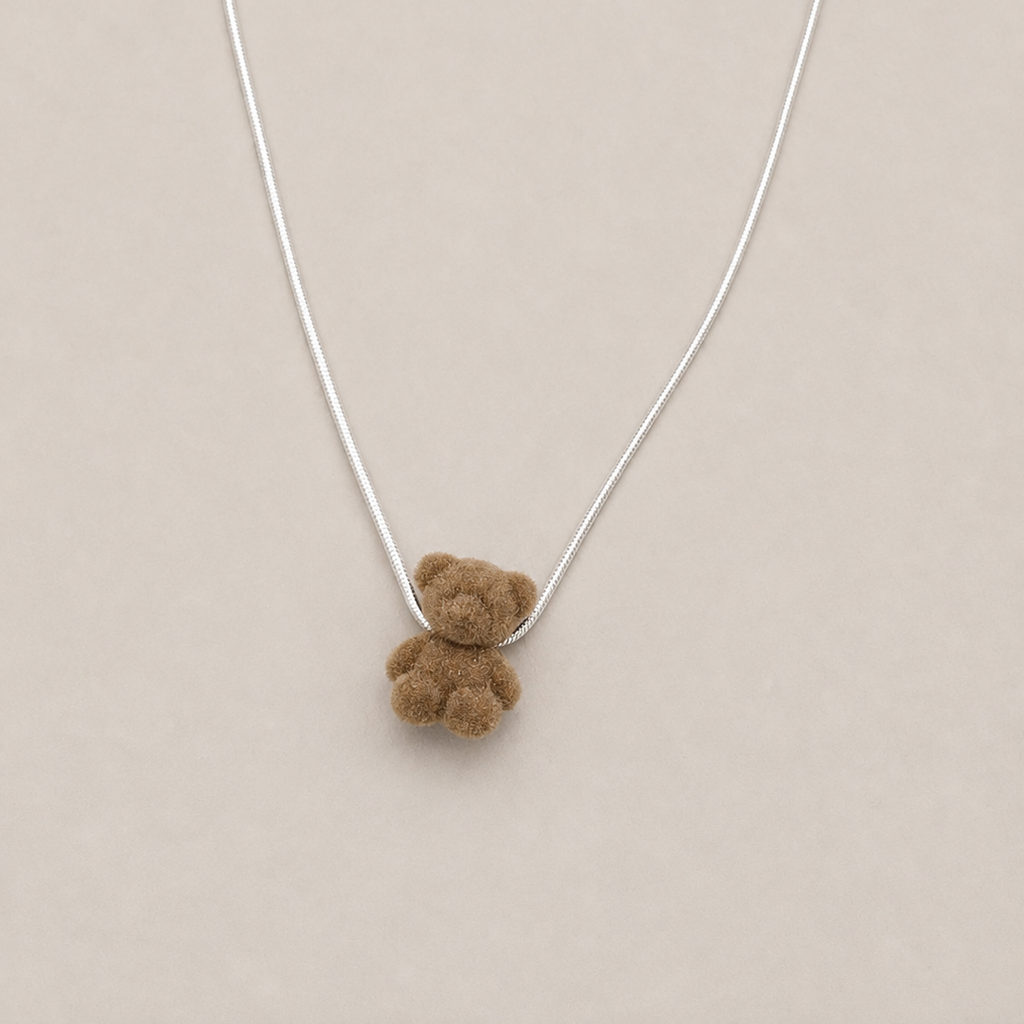 A silver chain necklace featuring a small, soft brown teddy bear pendant, displayed on a neutral beige background.