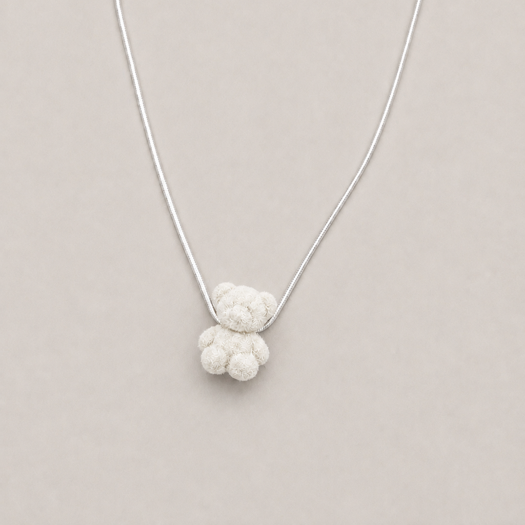 A delicate silver chain necklace featuring a small, textured white knotted pendant shaped like a teddy bear, presented on a solid light beige background.