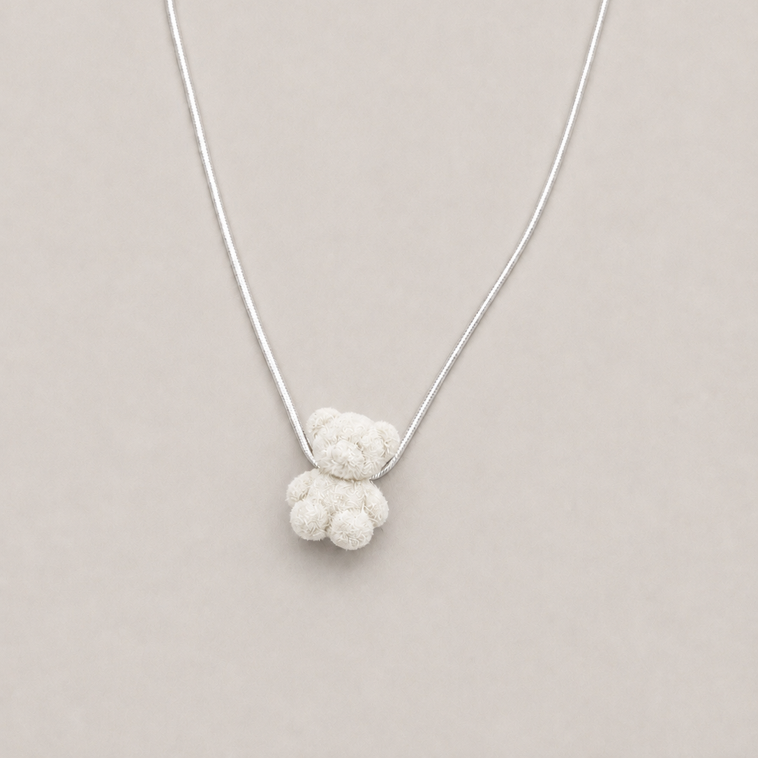 A delicate silver chain necklace featuring a small, textured white knotted pendant shaped like a teddy bear, presented on a solid light beige background.