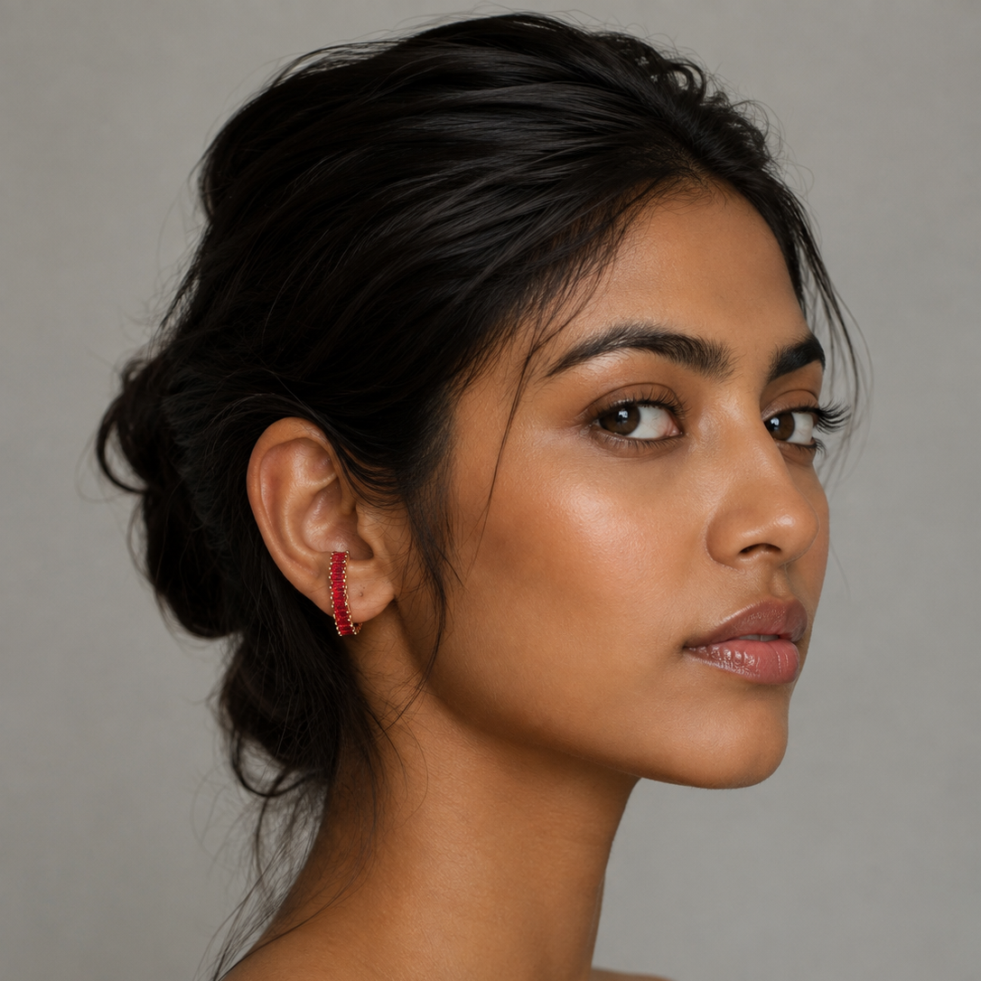 A close-up portrait of a woman with dark hair in an updo, wearing a small red geometric drop earring, set against a plain gray studio background.
