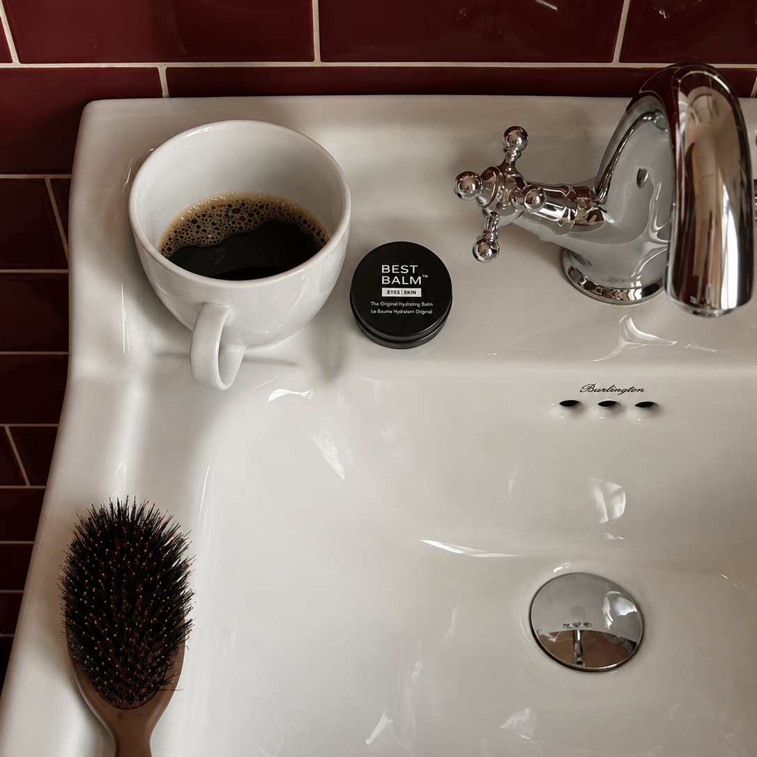 A white ceramic bathroom sink with a chrome faucet, a white mug filled with coffee, a small black jar labeled BEST BALM, and a brown-handled hairbrush resting on the sink. Dark red tiled walls are visible in the background.