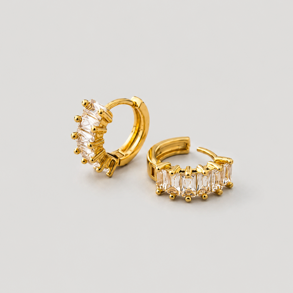 A pair of gold huggie hoop earrings, one featuring a cluster of clear baguette-cut stones and the other showing a side view with a row of matching clear stones, set against a plain light gray background.