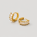 A pair of gold huggie hoop earrings, one featuring a cluster of clear baguette-cut stones and the other showing a side view with a row of matching clear stones, set against a plain light gray background.