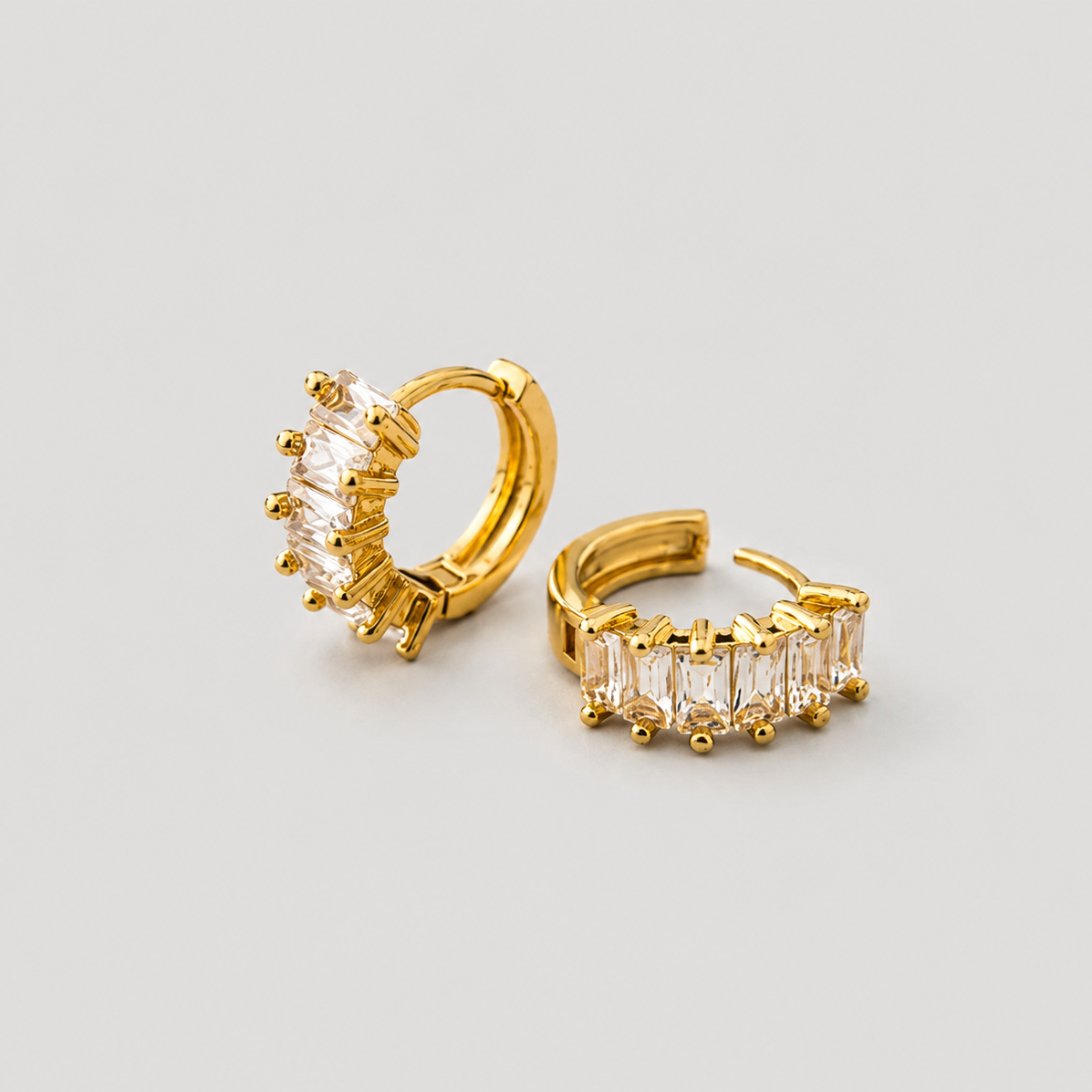 A pair of gold huggie hoop earrings, one featuring a cluster of clear baguette-cut stones and the other showing a side view with a row of matching clear stones, set against a plain light gray background.