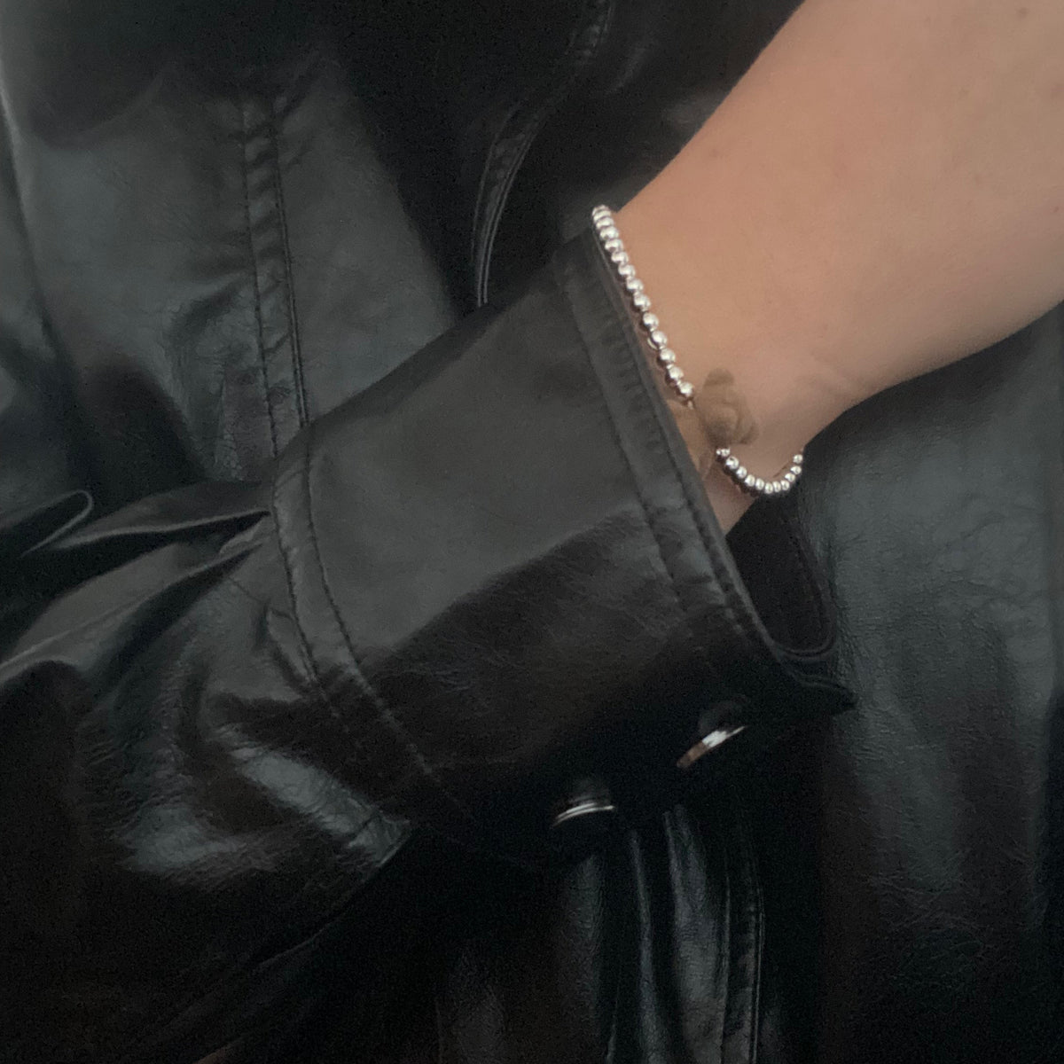 A close-up of a person wearing a silver beaded bracelet on their wrist, paired with a black faux-leather jacket and blue jeans.