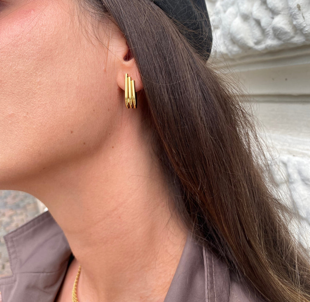 A close-up of a person's ear wearing a small gold earring with three parallel horizontal bars, next to long brown hair and a dark cap.