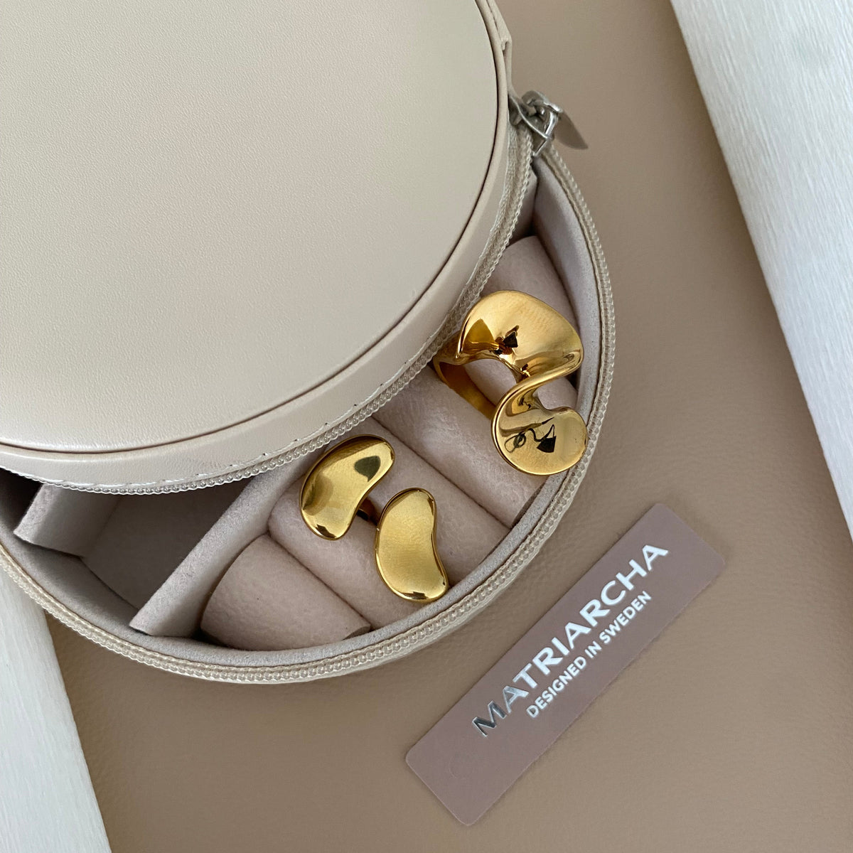 Gold bird-shaped stud earrings displayed in a round beige jewelry case, with a brand tag reading MATRARCHA placed beside them on a beige surface.