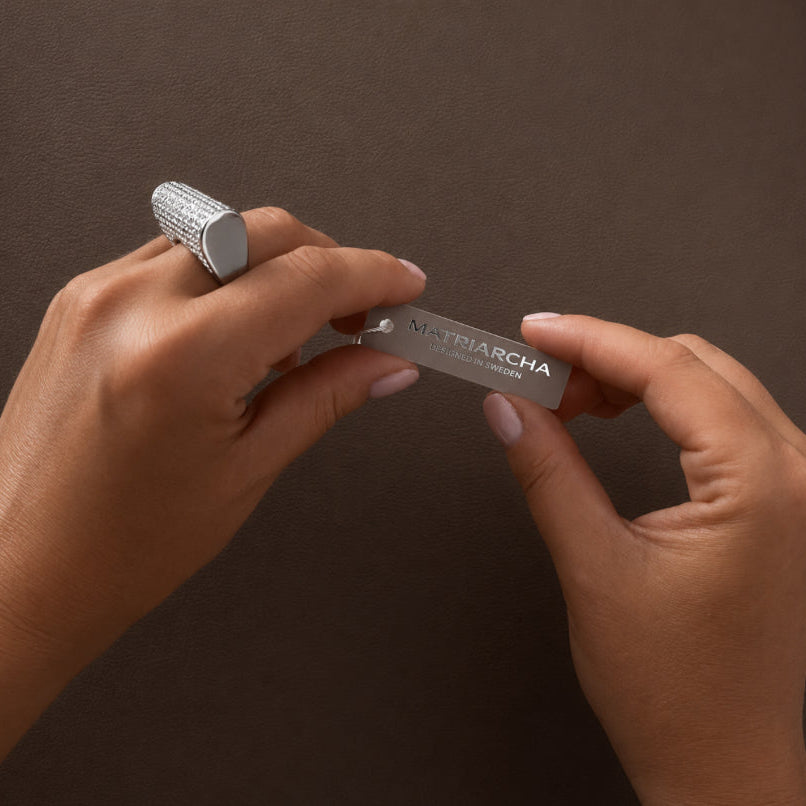 A close-up of a person's hands holding a slim silver metal nail file with the brand name 'MATRIARCHA' printed on it, against a dark brown textured background.