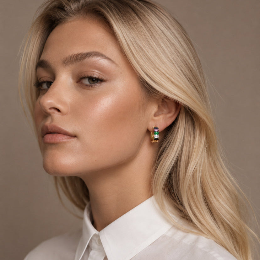 A close-up portrait of a blonde woman wearing a small gold hoop earring with green and black rectangular stones, paired with a white collared blouse against a neutral beige background.