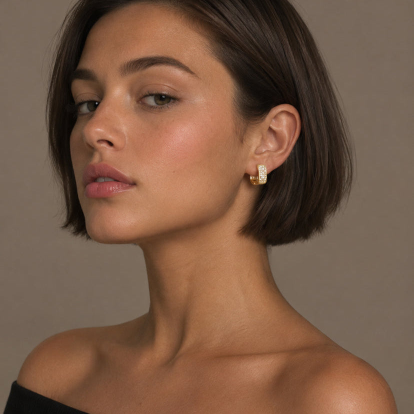 A woman with a short dark brown bob hairstyle wearing a small gold hoop earring with a white rectangular inlay, shown against a plain taupe background.
