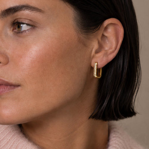 A close-up of a woman with dark hair wearing a delicate gold huggie hoop earring and a light pink knit sweater, set against a neutral beige background.