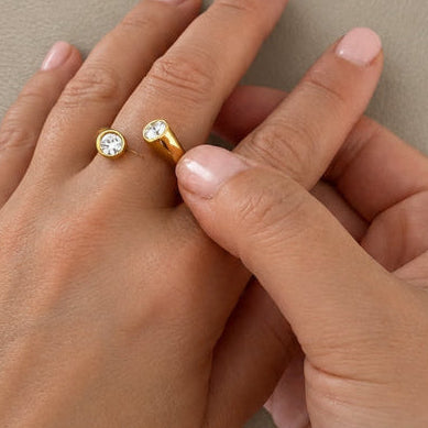 A close-up of two hands resting against a neutral beige background, with one hand wearing a delicate gold ring with a small clear gemstone.