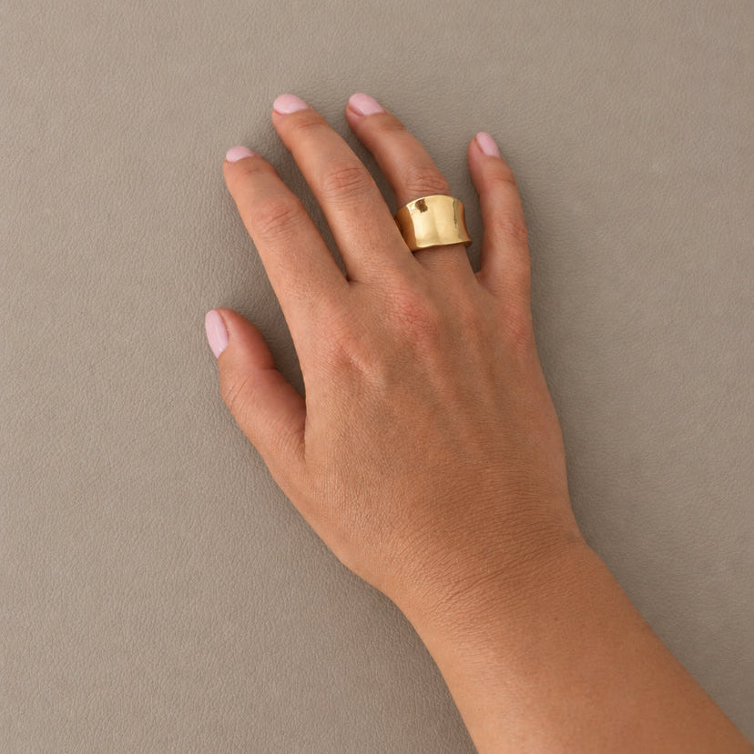 A gold ring with a small dark square detail on one of the fingers, shown on a model's hand against a neutral taupe background.