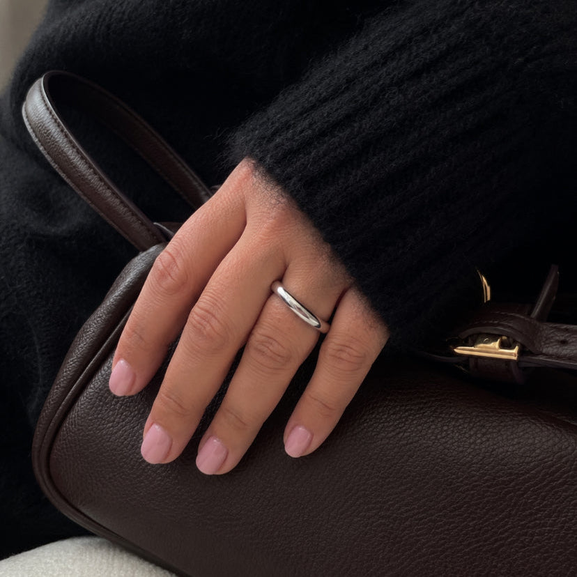A close-up of a hand with light pink manicured nails resting on a dark brown textured leather handbag, wearing a simple silver band ring. The person is dressed in a black fuzzy knit sweater.