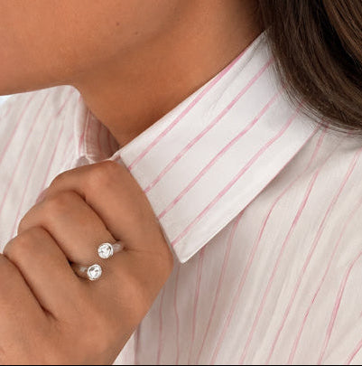A woman in profile wearing a white button-down shirt with thin pink stripes, featuring a small white rectangular stud earring and a silver ring with clear stones.