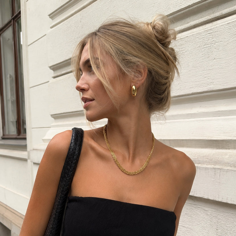 A woman wearing a black strapless tube top and light blue jeans, standing outdoors against a white paneled wall. She is carrying a black shoulder bag and wearing gold jewelry.