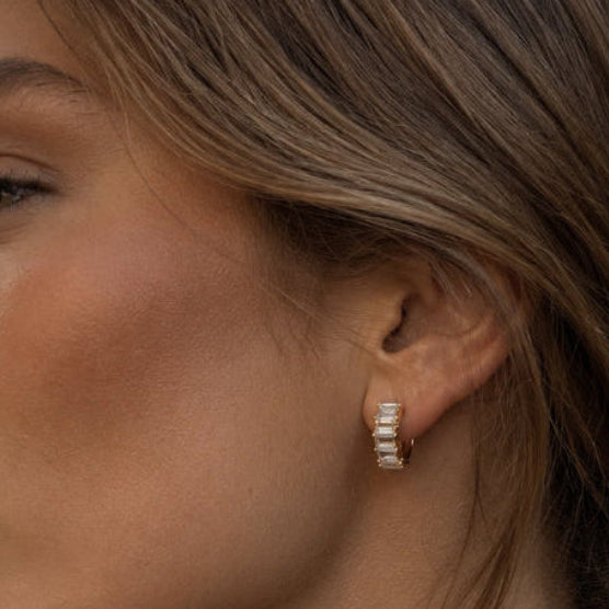 A close-up side profile of a woman with brown hair in a low bun, wearing a small silver-toned huggie hoop earring with clear stones, set against an outdoor urban background.