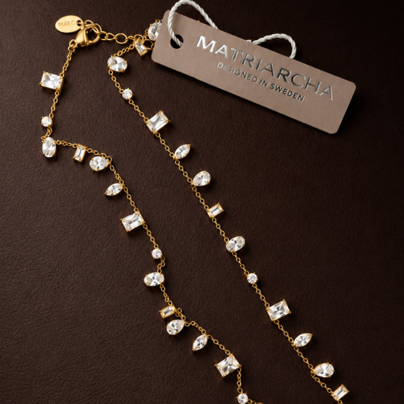 A gold-tone double-strand necklace with clear rhinestone and crystal drops, displayed on a dark brown background with a brand tag that reads 'MARIARICHA'.