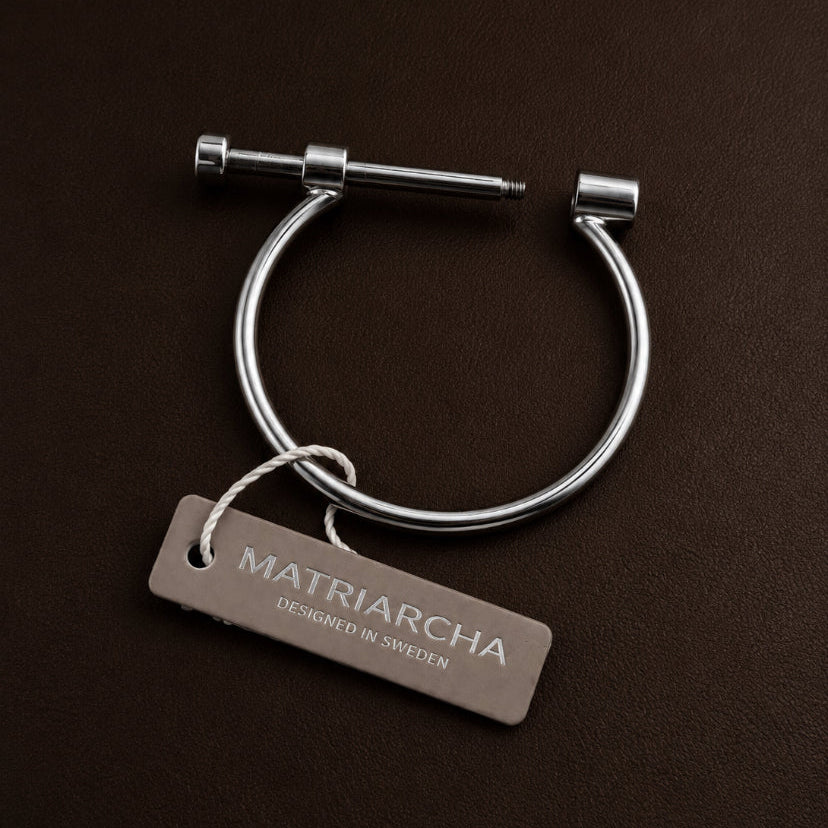 A silver-toned metal cuff bracelet with a sliding screw closure, shown on a dark brown textured background with a branded hangtag reading Matriarcha.