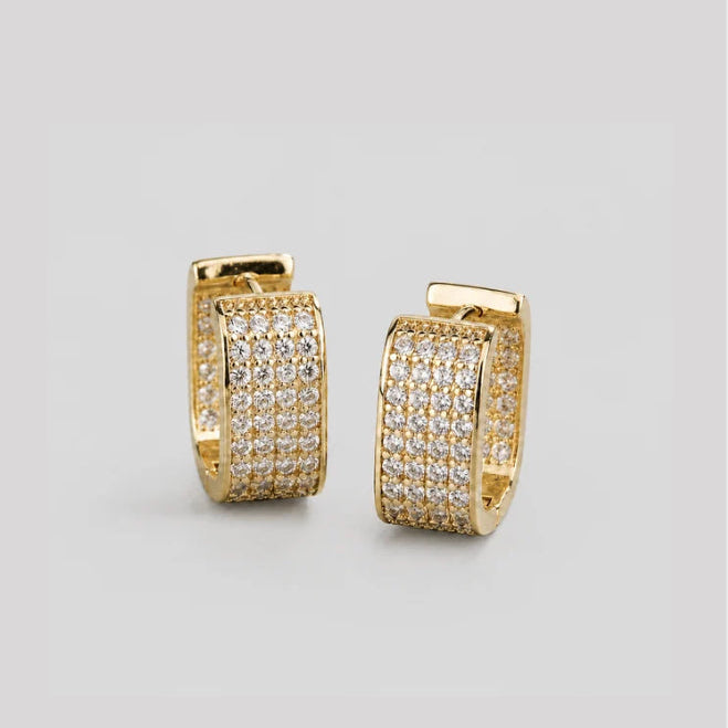A pair of gold huggie hoop earrings paved with small clear stones, displayed on a neutral light gray background.