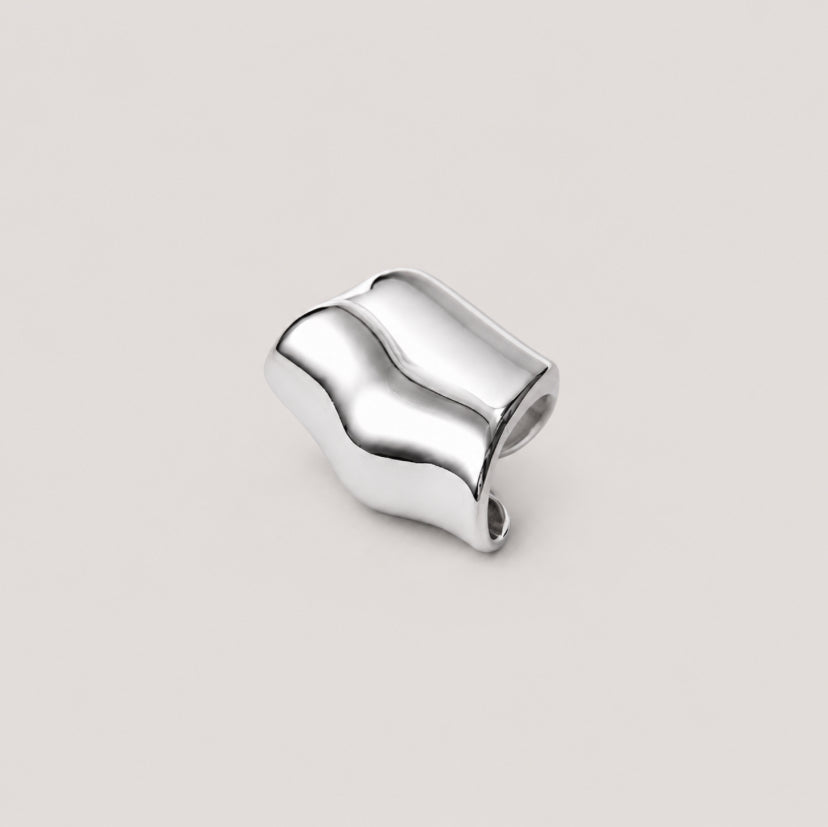 A polished silver earcuff with an organic, wavy, sculptural shape, photographed on a solid light beige background.