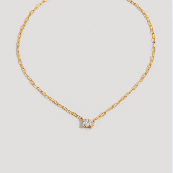 A delicate gold chain necklace with a small central gold bow-shaped setting holding a clear gemstone, displayed against a solid light gray background.