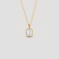A gold chain necklace with a bezel-set rectangular clear gemstone pendant, shown against a solid light gray background.