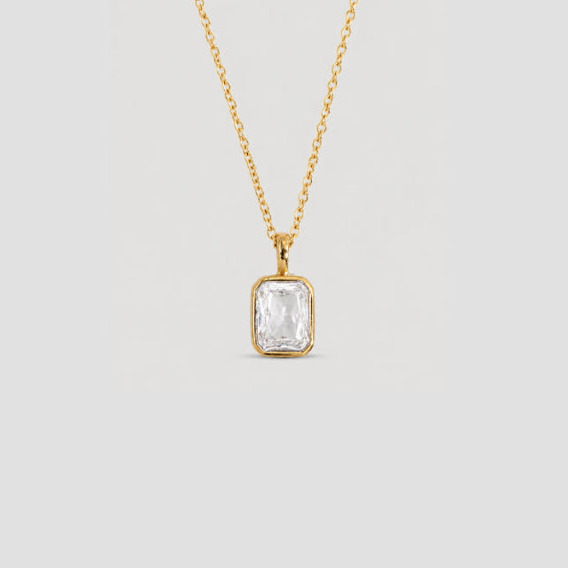 A gold chain necklace with a bezel-set rectangular clear gemstone pendant, shown against a solid light gray background.