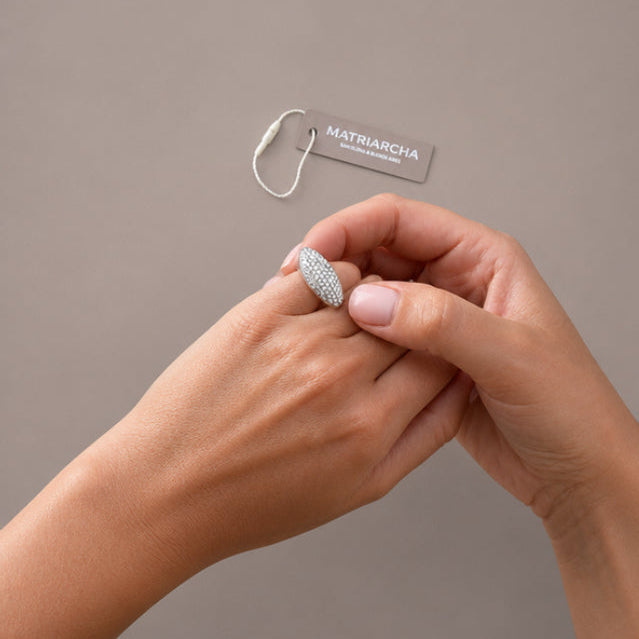 A close-up of a person's hands holding a silver ring with a textured, diamond-like pattern. A small brand tag for 'Matriarcha' is visible in the background against a solid taupe-gray backdrop.
