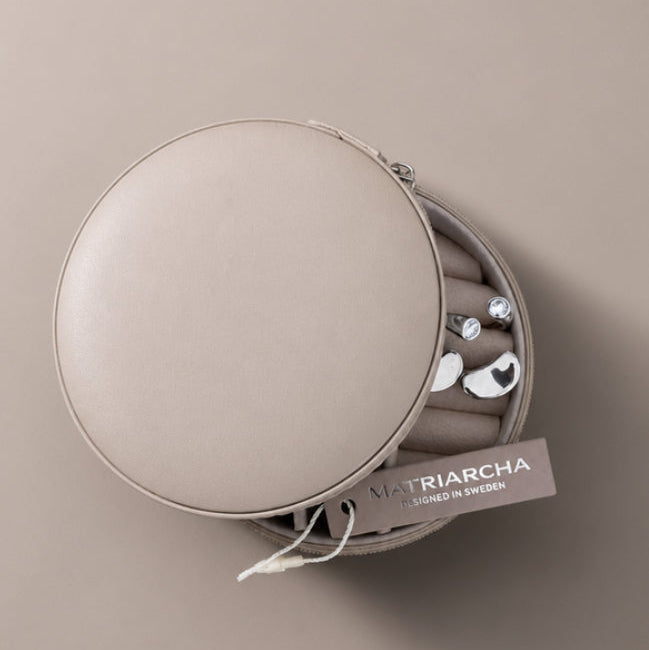 A round beige leather jewelry organizer case, partially open to reveal silver-toned jewelry inside, with a hanging tag reading Matriarcha on a matching beige background.