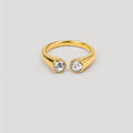 A gold open-front ring with two bezel-set clear round gemstones, shown on a light gray background.