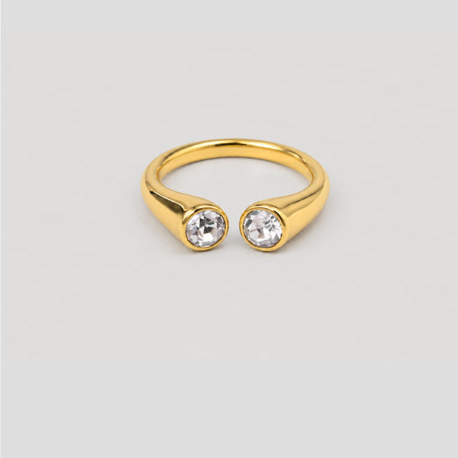 A gold open-front ring with two bezel-set clear round gemstones, shown on a light gray background.