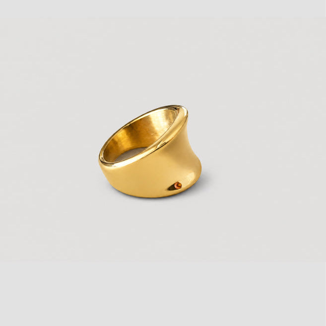 A gold ring with a wide, sculptural, tapered band, photographed on a neutral light gray background.