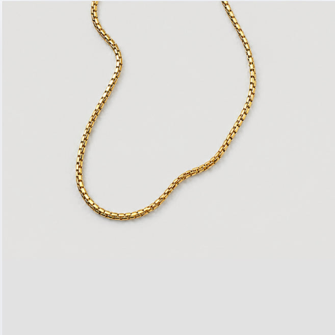 A gold-toned chain necklace with a textured, box-like link design, shown draped against a solid light gray background.