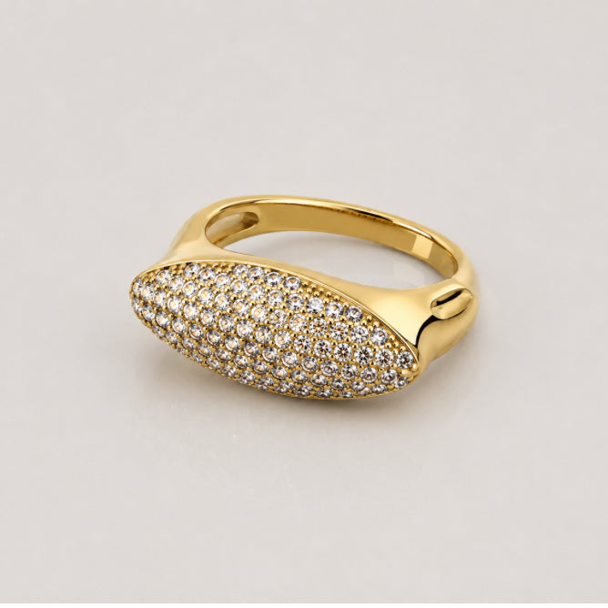 A gold ring featuring an elongated oval pavé-set bezel with clear stones, shown on a neutral light gray background.