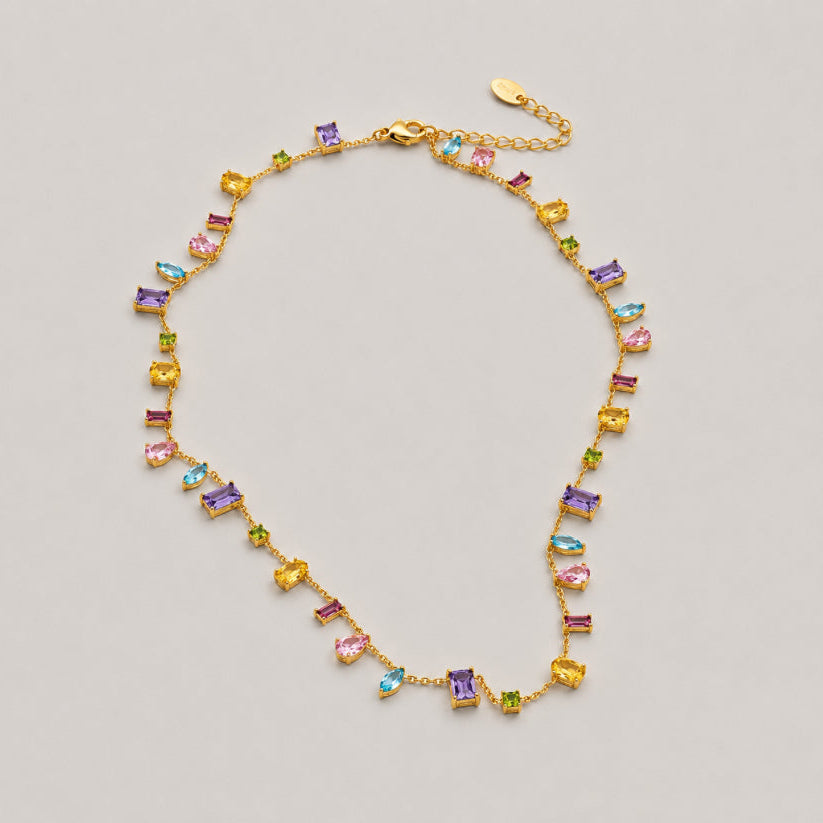 A gold chain necklace featuring multiple small rectangular gemstones in yellow, pink, purple, and green, arranged in a loose circle on a light gray background.