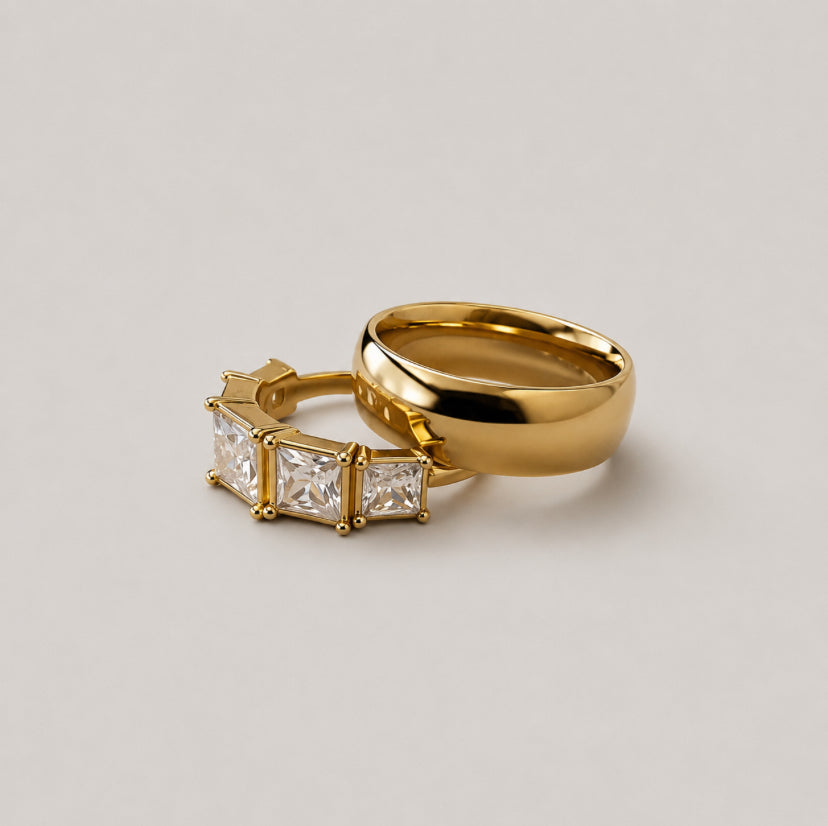 Two gold rings on a light gray background: one is a smooth, polished band ring, and the other is a ring featuring three princess-cut clear stones in a gold setting.