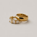 Two gold rings on a light gray background: one is a smooth, polished band ring, and the other is a ring featuring three princess-cut clear stones in a gold setting.