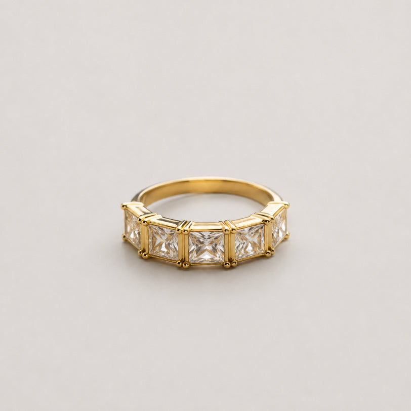 A yellow gold ring featuring a row of princess-cut clear gemstones in a bezel setting, photographed on a neutral light gray background.